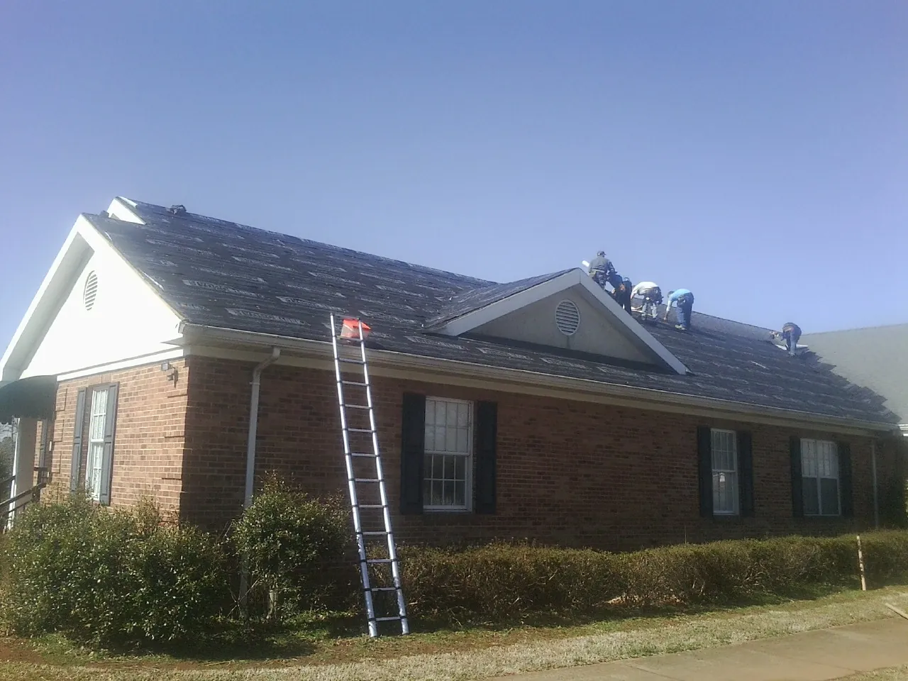 Crew tearing shingles off a brick ranch roof with a ladder set — Emergency Roof Repair work by our crew in East Lynn