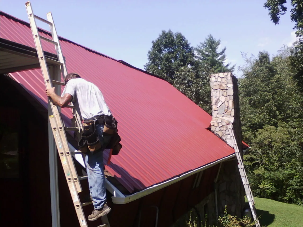 Roofer on a ladder beside a red metal roof and stone chimney — Soffit & Fascia Repair by Roofing Brothers in East Lynn